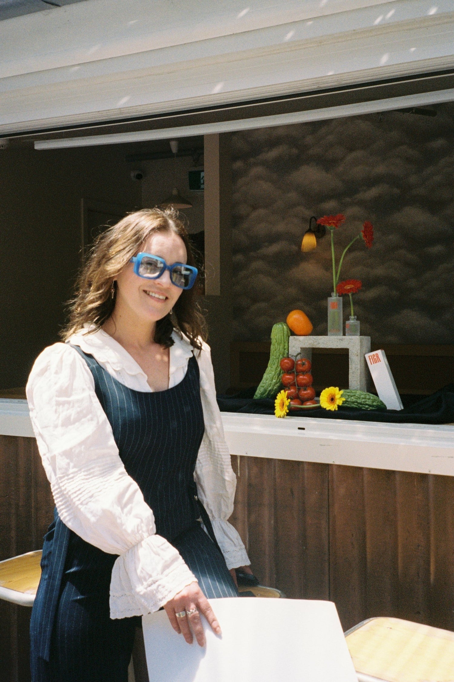 Melbourne based artist, still life club host and photographer Jay-Dee Pitcaithly wearing sunglasses standing in front of a window with decorative items.