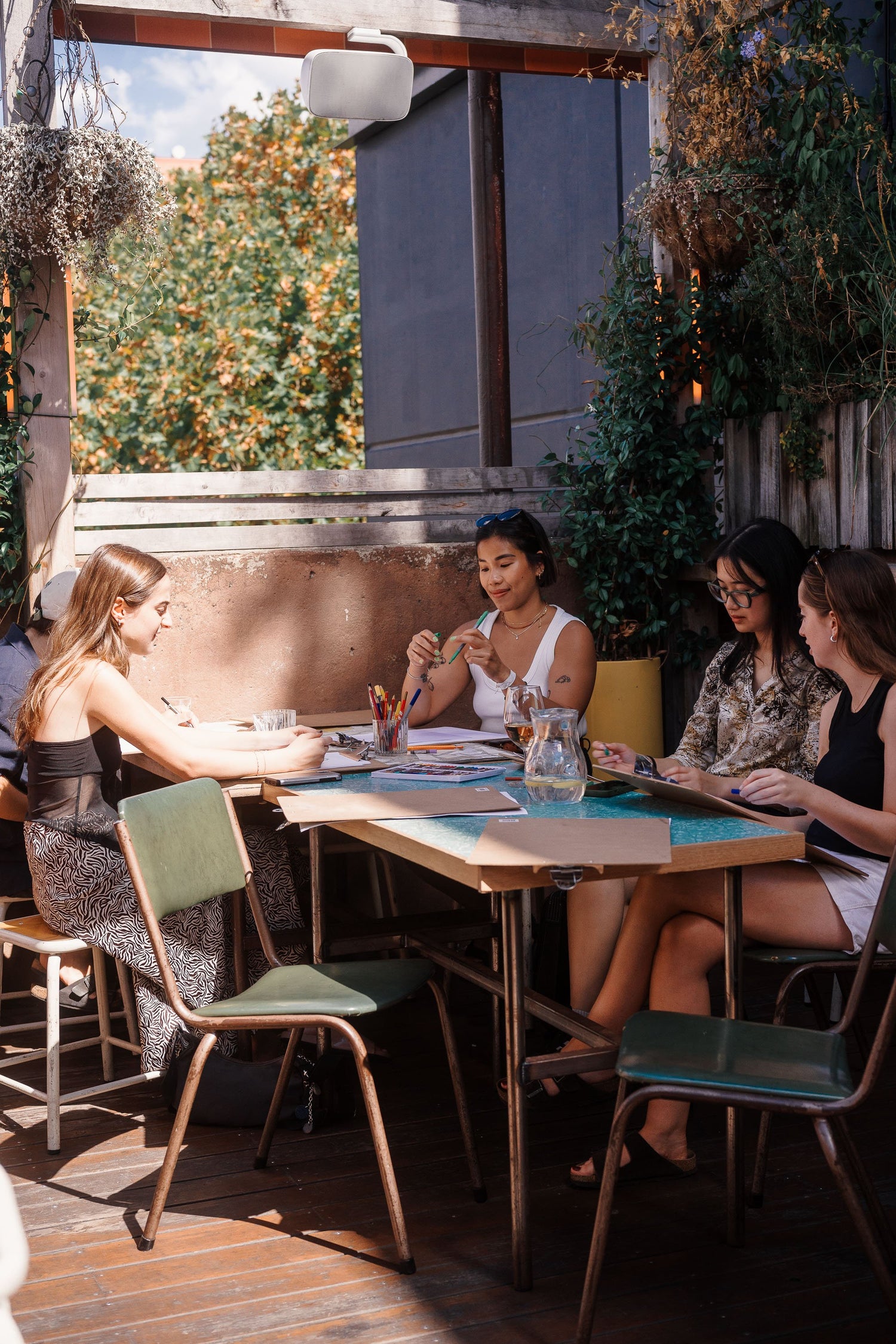 Group of people sitting at a table with plants and outdoor furniture drawing still life scenes on clipboards